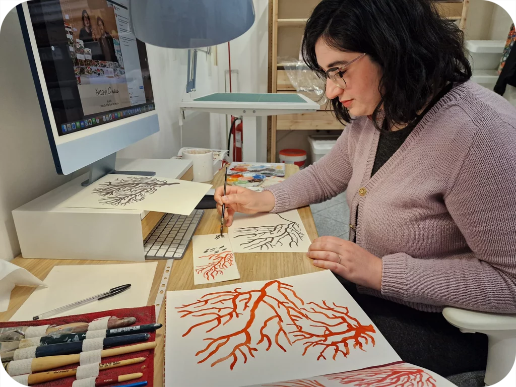 Eleonora Spada è di profilo seduta ad una scrivania mentre dipinge dei coralli stilizzati con un pennello su fogli di carta cotone all'interno del laboratorio di Versicò