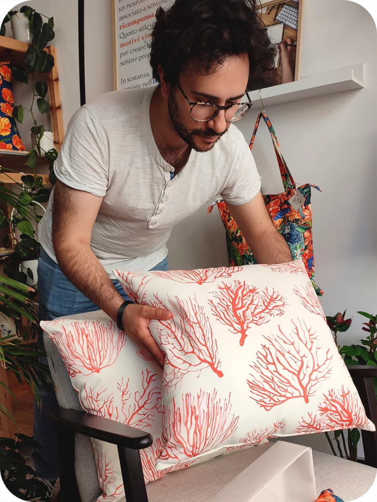 Uomo appoggia un cuscino in tessuto stampato con coralli rossi su fondo chiaro sopra una poltrona