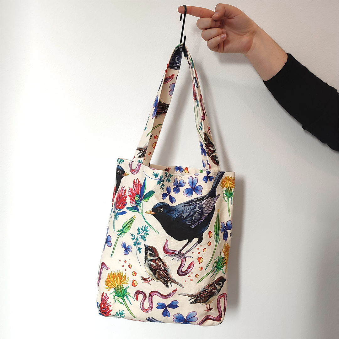 Una mano tiene un gancio a cui è appesa una borsa in tessuto stampato con un motivo con merli, passerotti, lombrichi, fiori e foglie su sfondo chiaro