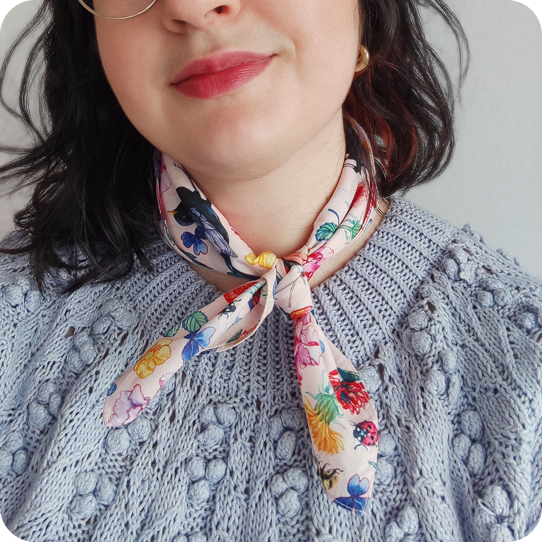 Dettaglio del collo di una donna che indossa bandana legata al collo realizzata con un tessuto in cotone stampato con chiocciole, api, coccinelle, merli, fragole, fiori e foglie su fondo rosa