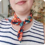 Dettaglio del collo di una donna che indossa una bandana legata al collo realizzata con un tessuto in cotone stampato con fiori di papavero, oleandro e bouganville arancioni, fiori di gelsomino bianchi e foglie verde-blu
