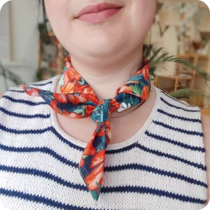 Dettaglio del collo di una donna che indossa una bandana legata al collo realizzata con un tessuto in cotone stampato con fiori di papavero, oleandro e bouganville arancioni, fiori di gelsomino bianchi e foglie verde-blu