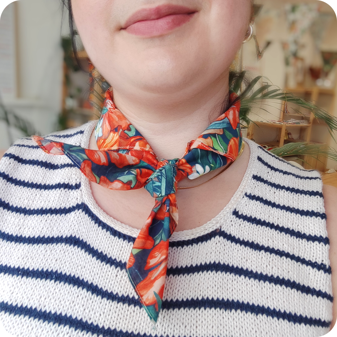 Dettaglio del collo di una donna che indossa una bandana legata al collo realizzata con un tessuto in cotone stampato con fiori di papavero, oleandro e bouganville arancioni, fiori di gelsomino bianchi e foglie verde-blu