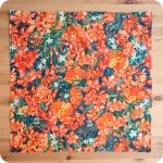 Bandana quadrata stesa sopra un tavolo in legno, realizzata con un tessuto in cotone stampato con fiori di papavero, oleandro e bouganville arancioni, fiori di gelsomino bianchi e foglie verde-blu
