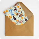 Biglietto d'auguri con gatti che saltano, biciclette, cappelletti, coppie di pane e statua del Savonarola su fondo azzurro; con busta avana