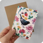 Biglietto d'auguri con un merlo al centro attorniato da fiori, foglie, coccinelle, api, farfalle e chiocciola su fondo rosa, con busta color avana