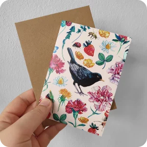 Biglietto d'auguri con un merlo al centro attorniato da fiori, foglie, coccinelle, api, farfalle e chiocciola su fondo rosa, con busta color avana