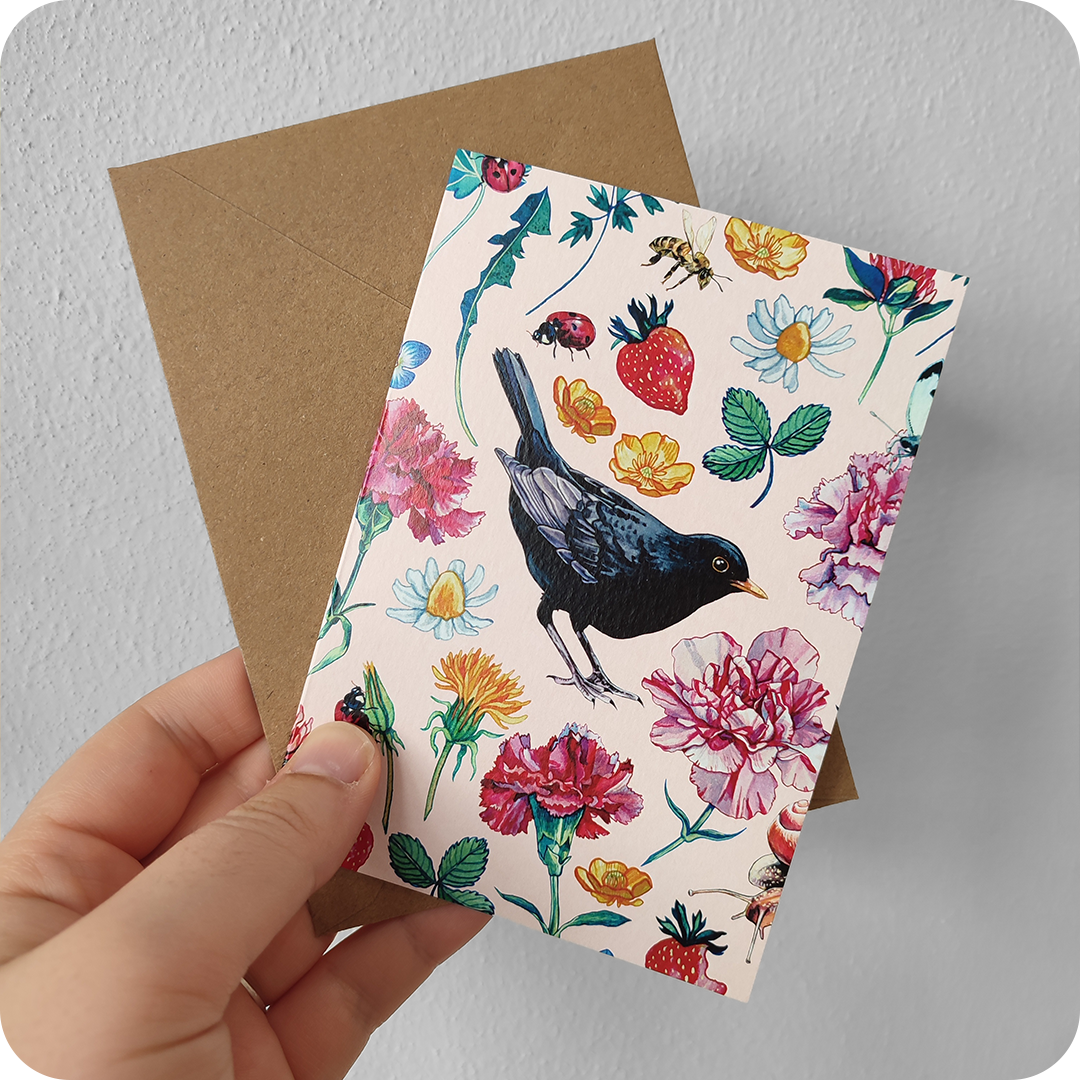 Biglietto d'auguri con un merlo al centro attorniato da fiori, foglie, coccinelle, api, farfalle e chiocciola su fondo rosa, con busta color avana