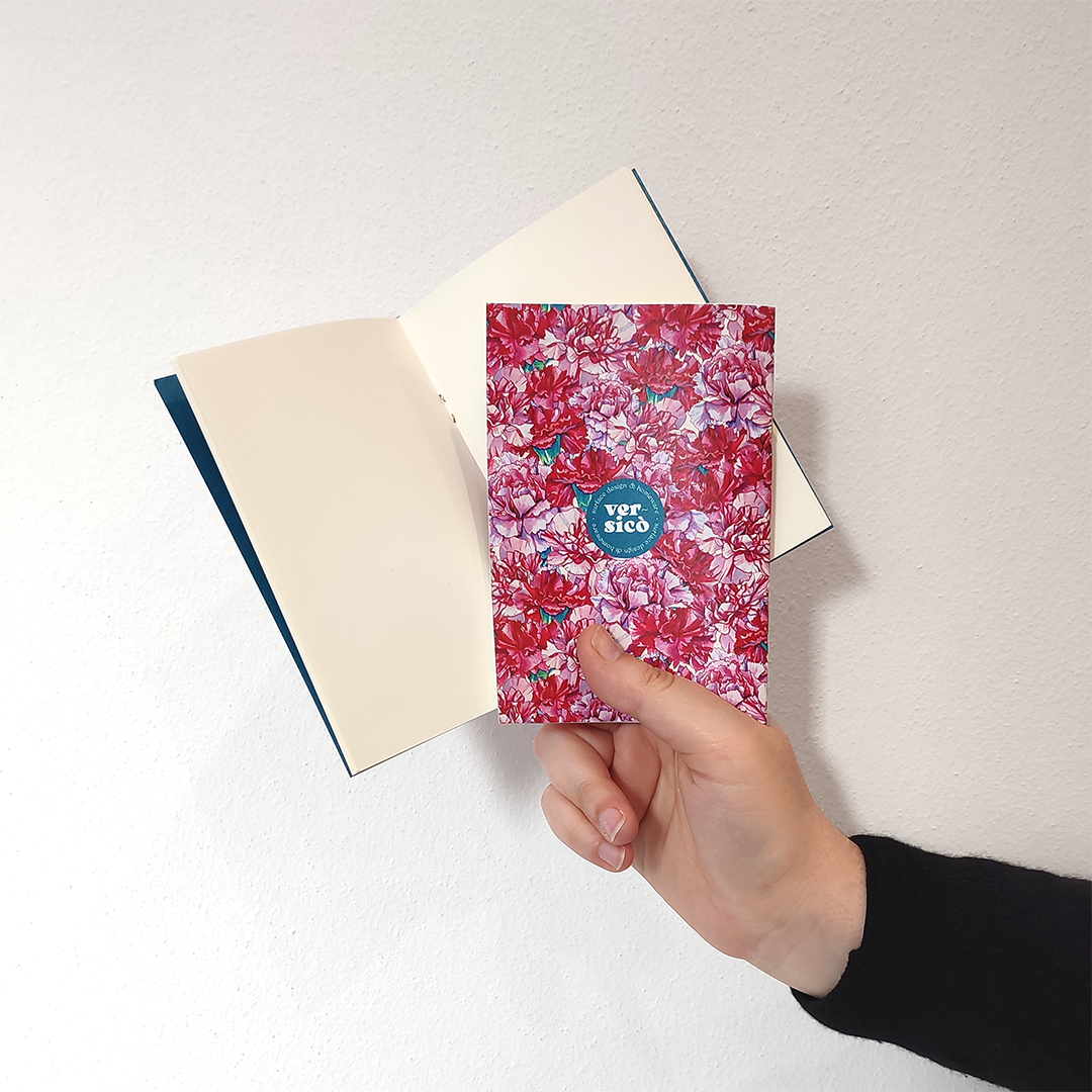 una mano mostra due taccuini, uno aperto con pagine bianche e uno chiuso con una copertina decorata con un pattern di garofani rosa e al centro il logo versicò