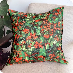 Dettaglio di un cuscino quadrato realizzato con un tessuto stampato con una fantasia con bacche e foglie appoggiato su una poltrona vista di tre quarti circondata da piante
