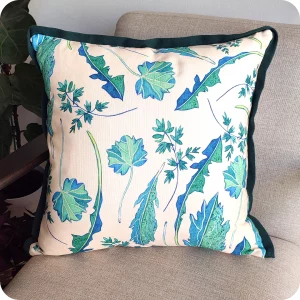 Dettaglio di un cuscino quadrato realizzato con un tessuto stampato con una fantasia con foglie verdi e azzurre su fondo chiaro appoggiato su una poltrona vista di tre quarti circondata da piante