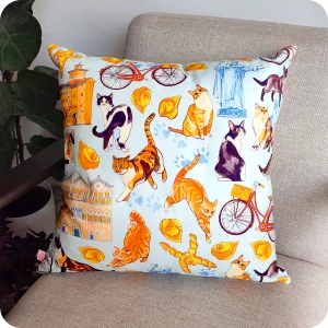 Dettaglio di cuscino quadrato realizzato con un tessuto stampato con una fantasia con gatti ed elementi legati a Ferrara appoggiato su una poltrona vista di tre quarti circondata da piante