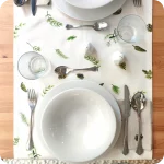 Runner apparecchiato realizzato con un tessuto stampato con una fantasia di rametti di pino, pigne e bacche su fondo bianco, con tovagliolo abbinato in lino écru