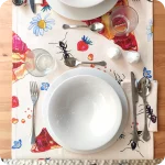 Runner apparecchiato realizzato con un tessuto stampato con disegni di fette di crostata, fiori, fragole e formiche su fondo chiaro, con tovagliolo abbinato