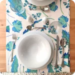 Runner apparecchiato realizzato con un tessuto stampato con disegni di foglie verdi e azzurre su fondo chiaro, con tovagliolo abbinato