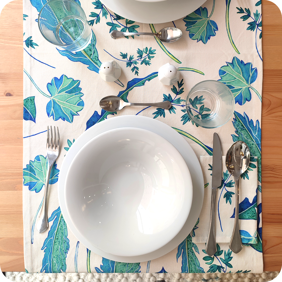 Runner apparecchiato realizzato con un tessuto stampato con disegni di foglie verdi e azzurre su fondo chiaro, con tovagliolo abbinato