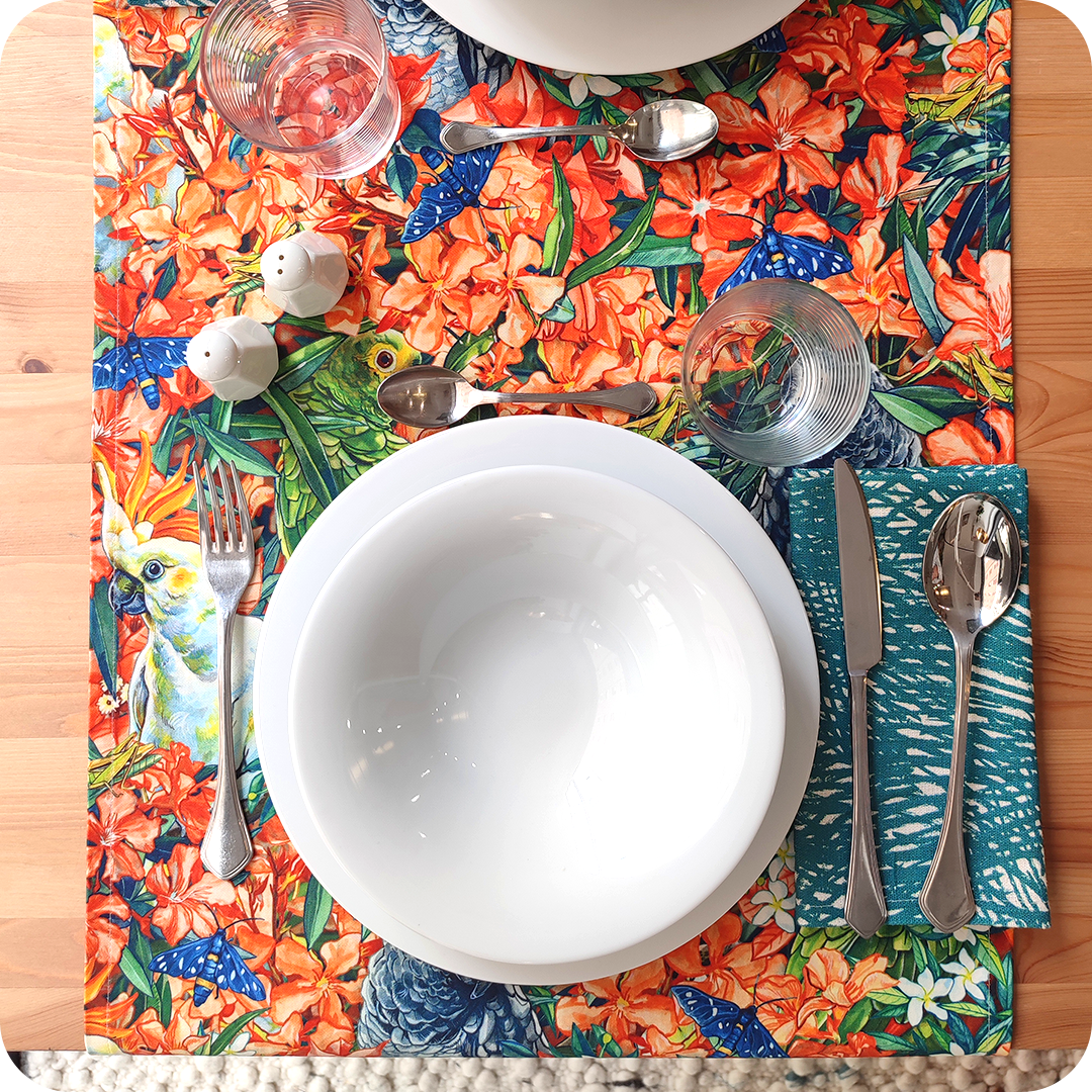 Runner apparecchiato realizzato con un tessuto stampato con pappagalli che sbucano da un fitto cespuglio fiorito su cui si poggiano qua e là grilli e falene, con tovagliolo abbinato bianco e verde