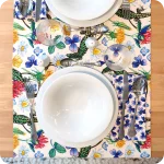 Runner apparecchiato in tessuto stampato con una fantasia con lucertole, fiori e foglie su fondo chiaro, tovagliolo in lino abbinato con trifogli blu su fondo chiaro