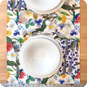 Runner apparecchiato in tessuto stampato con una fantasia con lucertole, fiori e foglie su fondo chiaro, tovagliolo in lino abbinato con trifogli blu su fondo chiaro