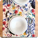 Runner apparecchiato in tessuto stampato con una fantasia con merli, passerotti, lombrichi, fiori e foglie su fondo chiaro, tovagliolo in lino abbinato con trifogli blu su fondo chiaro