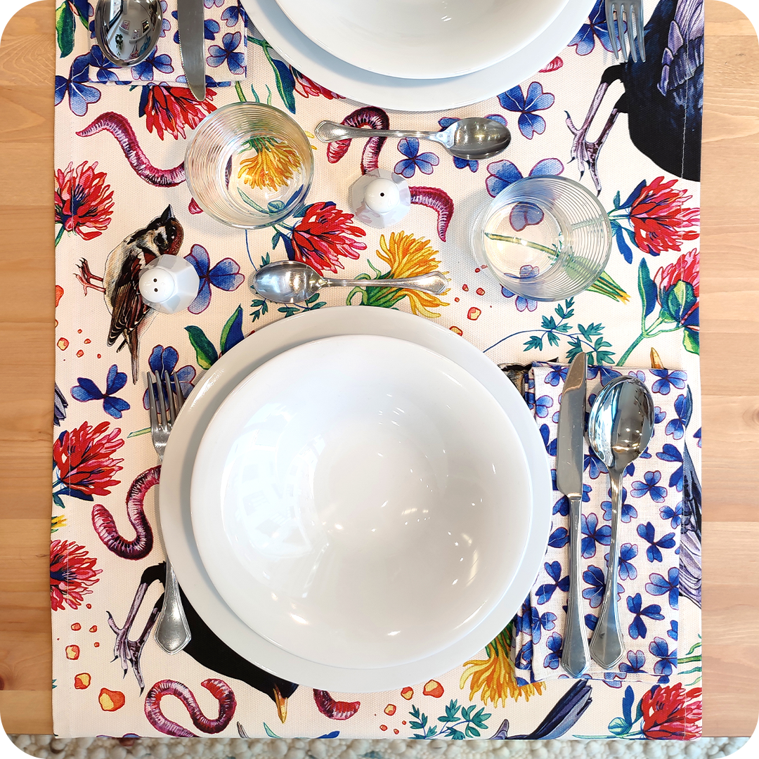 Runner apparecchiato in tessuto stampato con una fantasia con merli, passerotti, lombrichi, fiori e foglie su fondo chiaro, tovagliolo in lino abbinato con trifogli blu su fondo chiaro