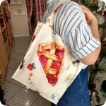Donna che indossa su una spalla una borsa in tessuto stampato con una fantasia con fette di crostata, formiche, fiori e fragole su fondo chiaro