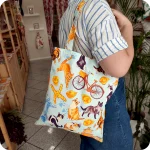 Donna che indossa su una spalla una borsa in tessuto stampato con una fantasia con gatti, cappelletti o tortellini, coppie di pane, biciclette e altri elementi legati a Ferrara, su sfondo azzurro