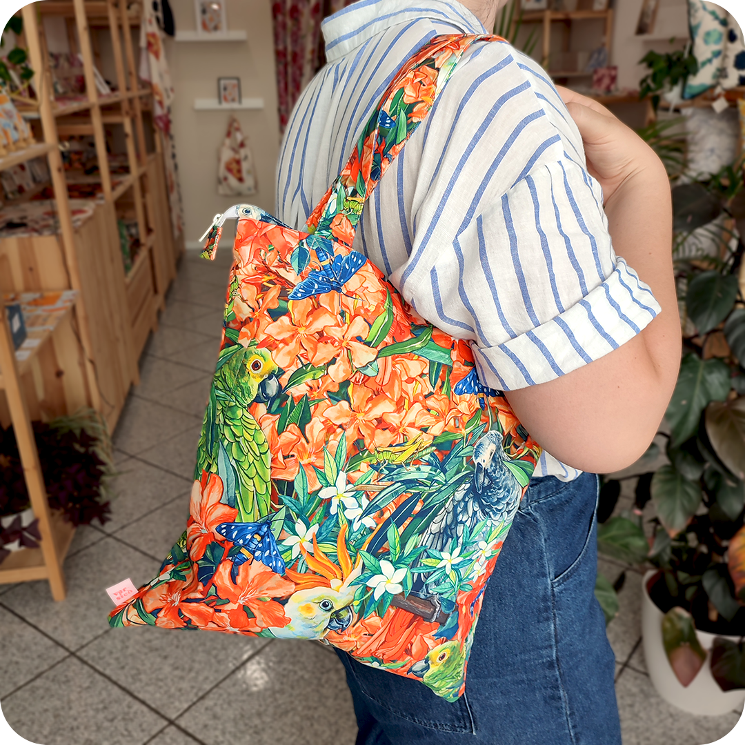 Donna che indossa su una spalla una borsa con cerniera in tessuto stampato con una fantasia jungle con pappagalli e falene che sbucano tra fiori e foglie