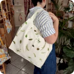 Donna che indossa su una spalla una grande borsa in tessuto stampato con un motivo con rametti di pino, bacche e pigne su fondo bianco