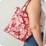 Donna che indossa su una spalla una borsa con cerniera in tessuto stampato con una fantasia con grappoli d'uva, rose canine e farfalle su fondo rosso scuro