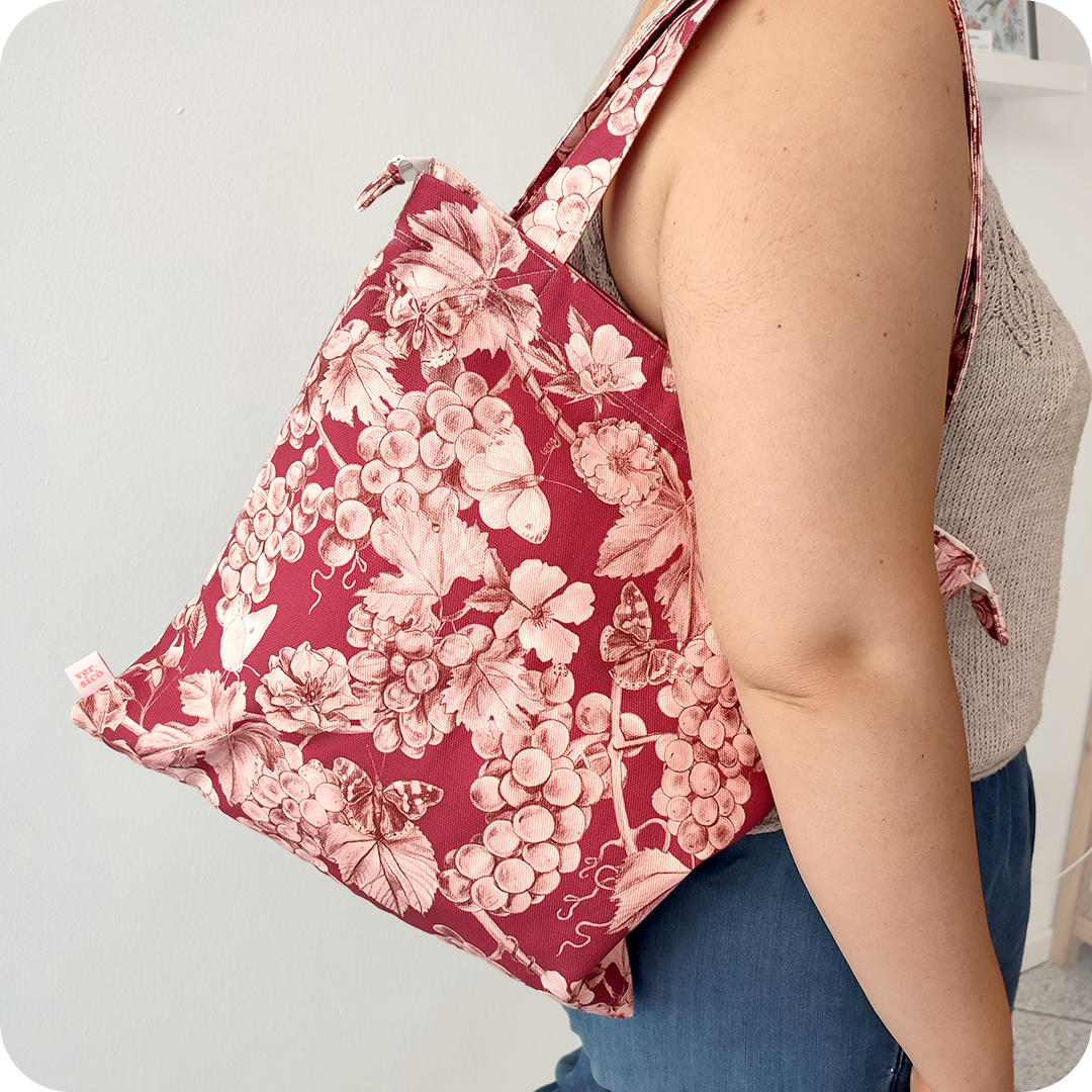 Donna che indossa su una spalla una borsa con cerniera in tessuto stampato con una fantasia con grappoli d'uva, rose canine e farfalle su fondo rosso scuro