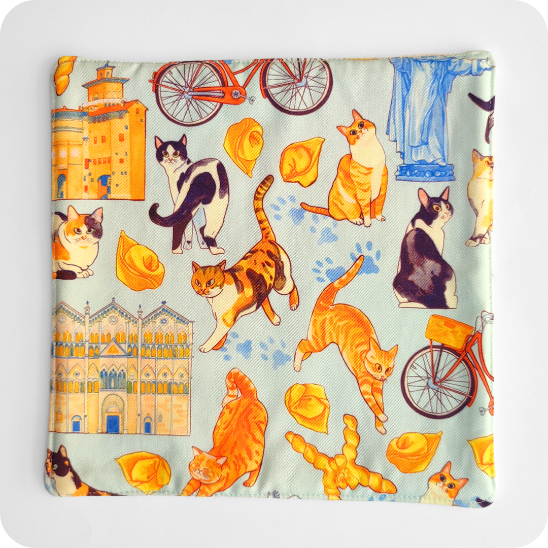 Sottopentola in tessuto imbottito realizzato con una stoffa stampata con gatti, cappelletti, coppie di pane, biciclette, duomo e castello di Ferrara su fondo azzurro