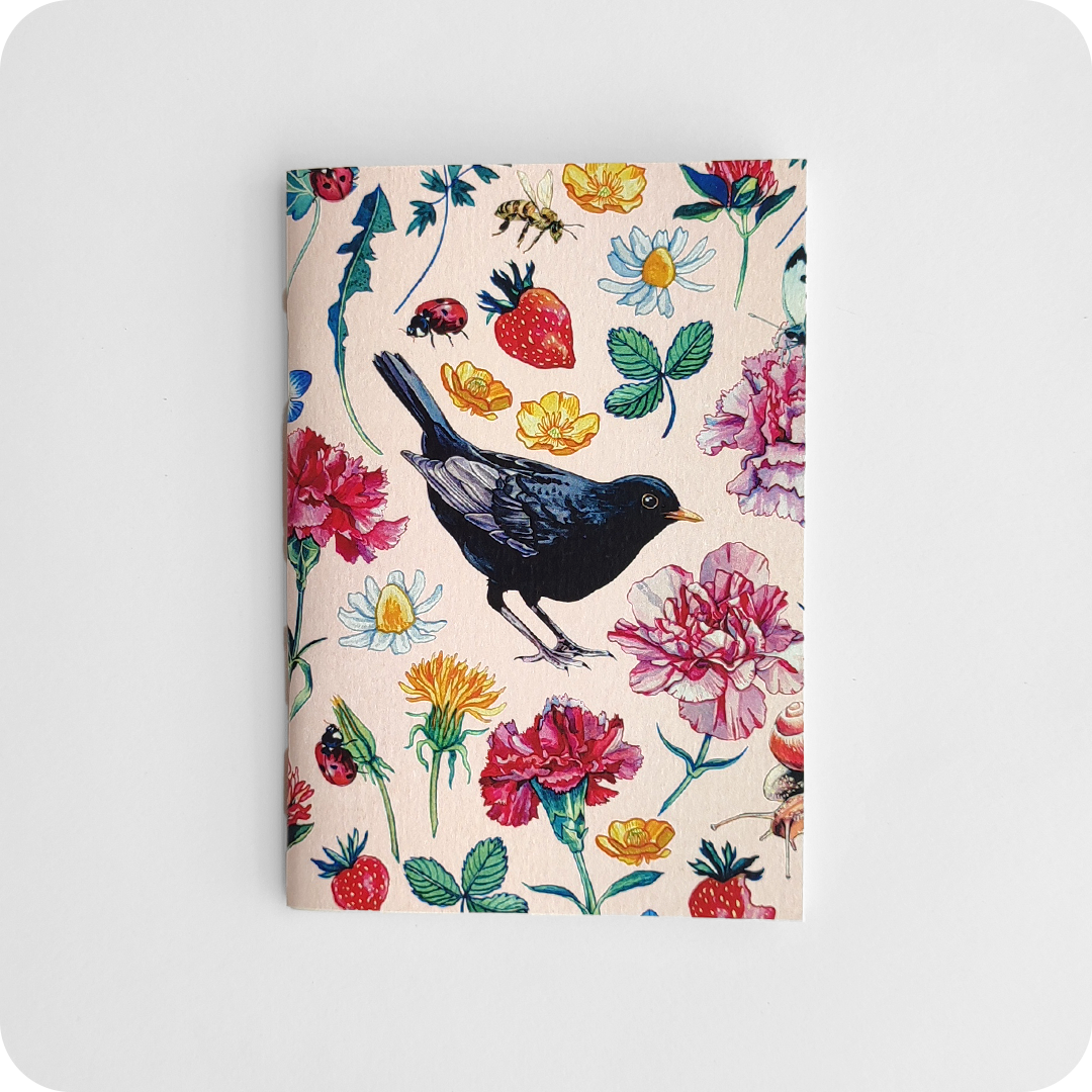 Fronte di un taccuino decorato con un merlo al centro attorniato da fiori, fragole, coccinelle, api su un fondo rosa