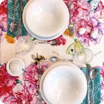 Tovaglia apparecchiata realizzata in tessuto stampato con festoni fiori, api e farfalle su fondo chiaro; tovagliolo abbinato con motivo a tratteggi verdi su fondo bianco