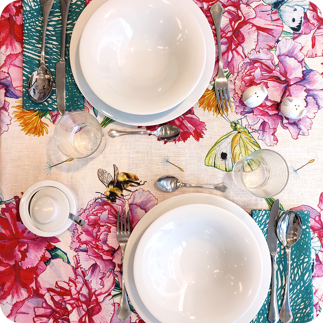 Tovaglia apparecchiata realizzata in tessuto stampato con festoni fiori, api e farfalle su fondo chiaro; tovagliolo abbinato con motivo a tratteggi verdi su fondo bianco