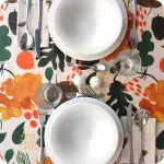 Tovaglia apparecchiata realizzata con un tessuto in lino naturale stampato con una fantasia con elementi vegetali stilizzati sui toni del giallo, arancione, verde e bruno