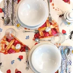 Tovaglia apparecchiata realizzata in tessuto stampato con fette di crostata, fragole, formiche e fiori su fondo chiaro; tovaglioli abbinati con tratteggi bianchi su fondo blu