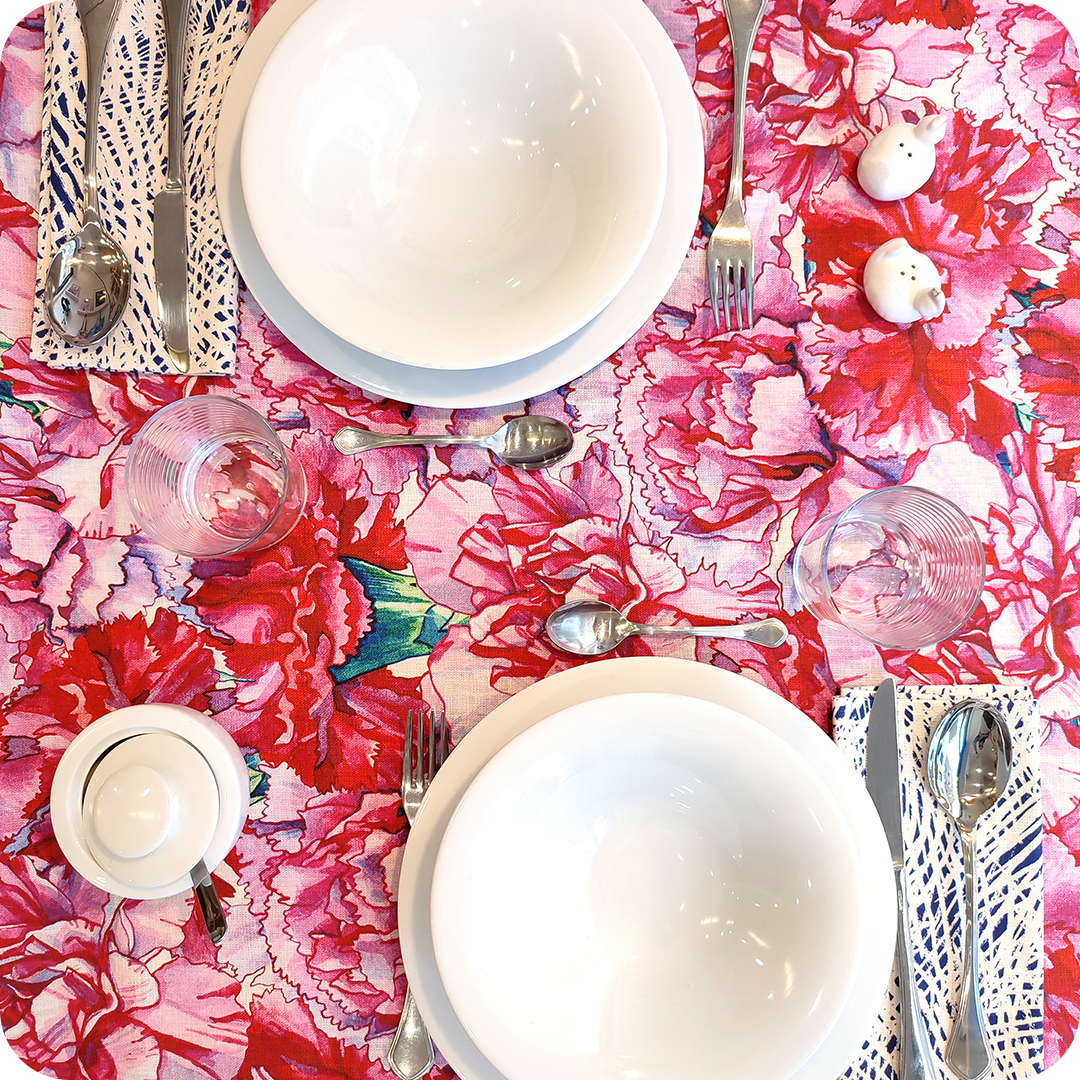 Tovaglia apparecchiata realizzata in tessuto stampato con garofani rosa e magenta; tovaglioli abbinati con motivo a tratteggi bianchi su fondo blu