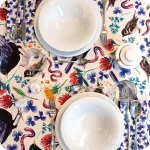 Tovaglia apparecchiata realizzata in tessuto stampato con trifogli blu, merli, passerotti, lombrichi, fiori di trifoglio rosso e tarassaco, foglie su fondo chiaro; tovaglioli abbinati con trifogli blu su fondo bianco