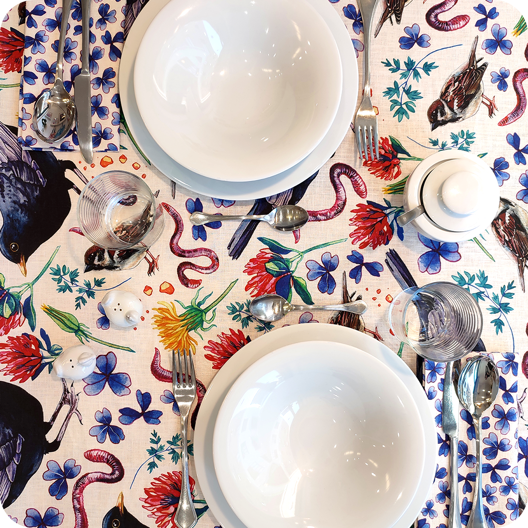Tovaglia apparecchiata realizzata in tessuto stampato con trifogli blu, merli, passerotti, lombrichi, fiori di trifoglio rosso e tarassaco, foglie su fondo chiaro; tovaglioli abbinati con trifogli blu su fondo bianco