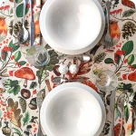 Tovaglia apparecchiata realizzata con un tessuto in lino naturale stampato con una fantasia con disegni di pettirossi, funghi, foglie, pigne, ghiande, fiori e bacche