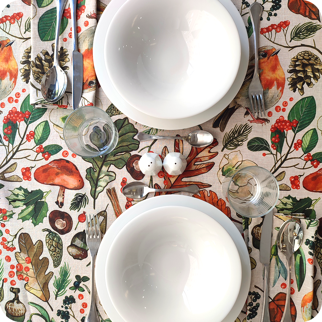 Tovaglia apparecchiata realizzata con un tessuto in lino naturale stampato con una fantasia con disegni di pettirossi, funghi, foglie, pigne, ghiande, fiori e bacche
