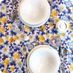 Tovaglia apparecchiata realizzata in tessuto stampato con trifogli blu, coccinelle, api, fiori gialli, azzurri e bianchi su fondo chiaro; tovaglioli abbinati con trifogli blu su fondo chiaro