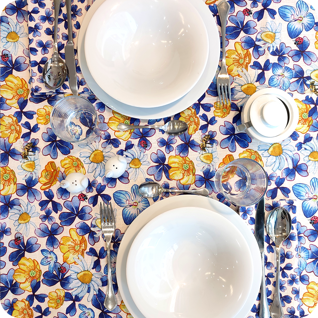 Tovaglia apparecchiata realizzata in tessuto stampato con trifogli blu, coccinelle, api, fiori gialli, azzurri e bianchi su fondo chiaro; tovaglioli abbinati con trifogli blu su fondo chiaro