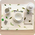 Tovaglietta rettangolare apparecchiata realizzata con un tessuto stampato con una fantasia con pigne e rametti di pino su fondo bianco; tovagliolo abbinato in lino naturale