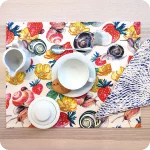 Tovaglietta rettangolare apparecchiata realizzata con un tessuto stampato con una fantasia con chiocciole, fiori e fragole su fondo chiaro; tovagliolo in lino abbinato con motivo a tratteggio bianco su fondo blu