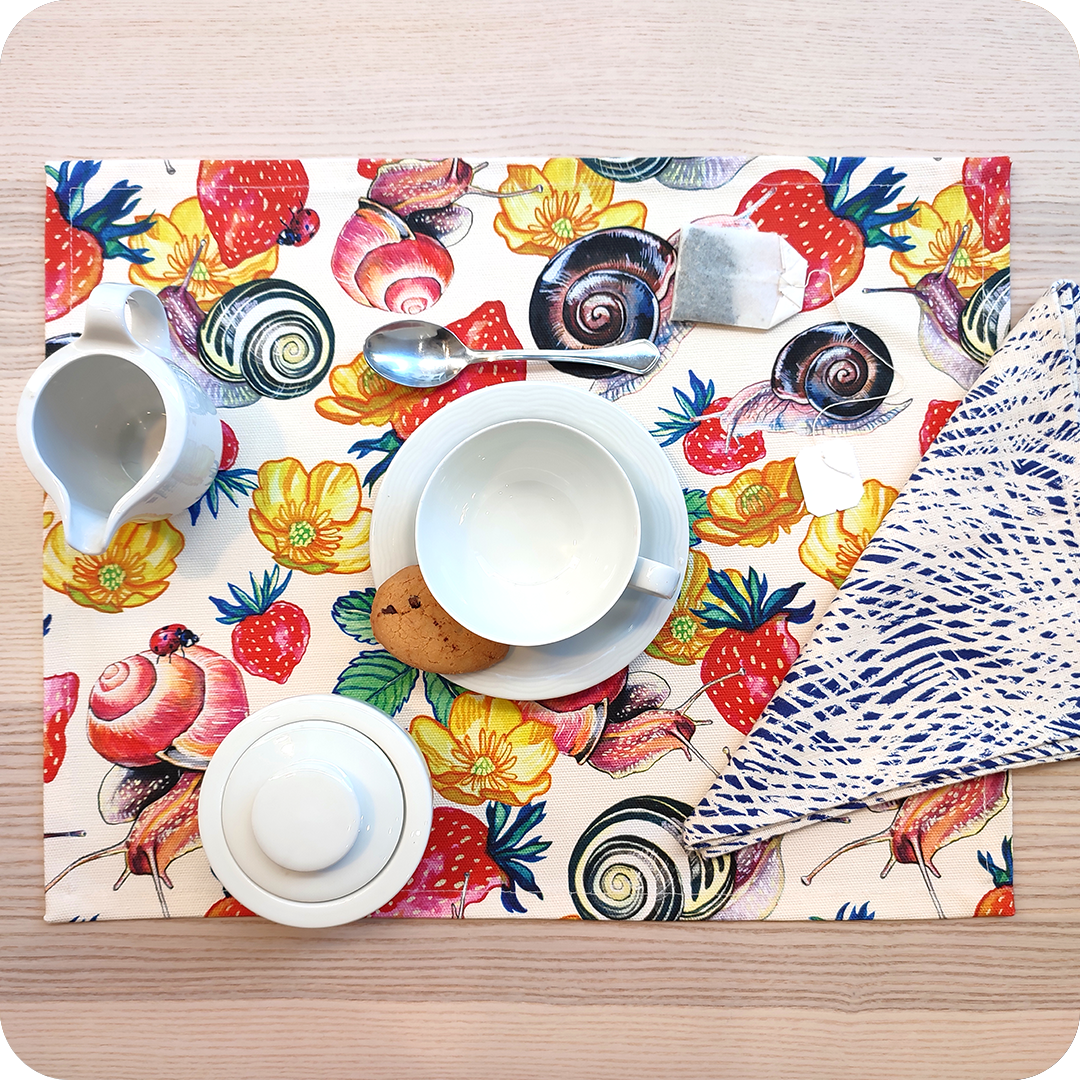 Tovaglietta rettangolare apparecchiata realizzata con un tessuto stampato con una fantasia con chiocciole, fiori e fragole su fondo chiaro; tovagliolo in lino abbinato con motivo a tratteggio bianco su fondo blu