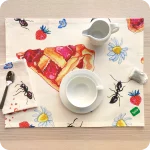 Tovaglietta rettangolare apparecchiata realizzata con un tessuto stampato con una fantasia con fette di crostata, fiori, formiche e fragole su fondo chiaro; tovagliolo in lino abbinato