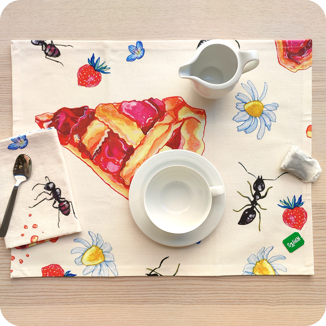 Tovaglietta rettangolare apparecchiata realizzata con un tessuto stampato con una fantasia con fette di crostata, fiori, formiche e fragole su fondo chiaro; tovagliolo in lino abbinato