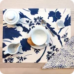 Tovaglietta rettangolare apparecchiata realizzata con un tessuto stampato con una fantasia di foglie blu su fondo chiaro; tovagliolo in lino abbinato con un motivo a tratteggi bianchi su fondo blu